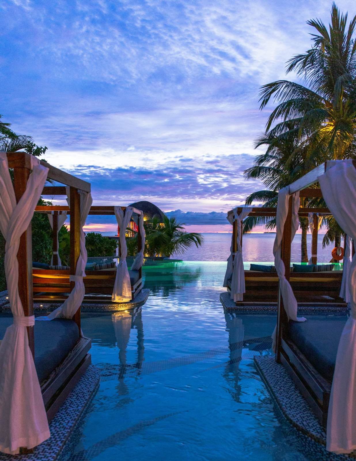 Balinese daybeds with white curtains flanking an infinity pool, facing the sea at dusk